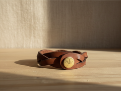 Hidden House Co. brown braided leather bracelet with a circular brass snap closure featuring a branded logo.