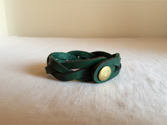 Hidden House Co. forest green braided leather wristband with an antique brass button closure on a white surface
