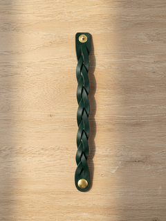 Hidden House Co. forest green braided leather wristband with antique brass button closures on a light wood surface.