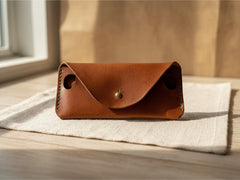 Hidden House Co. handcrafted brown leather glasses case with a brass stud closure and side cut-outs on a linen cloth.