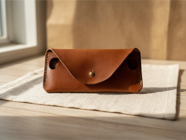 Hidden House Co. handcrafted brown leather glasses case with a brass stud closure and side cut-outs on a linen cloth.