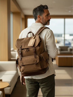 Hidden House Co. tan leather luggage tag attached to a brown canvas backpack worn by a man in an airport lounge