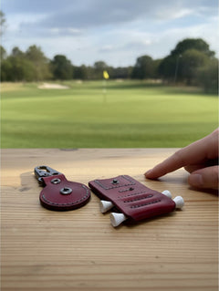 Hidden House Co. handcrafted deep red leather golf ball marker holder and tee caddy on a wooden table at a golf course