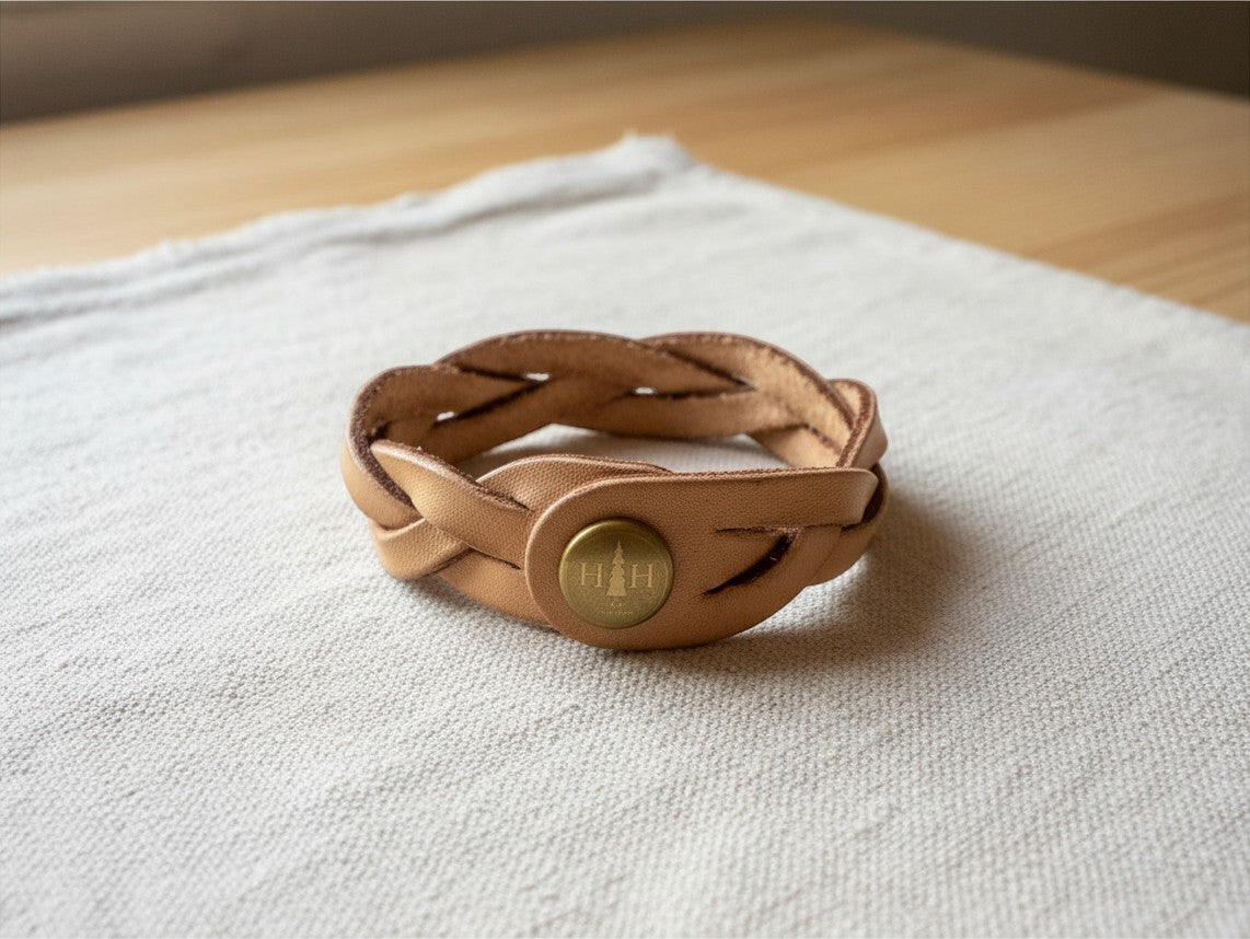 Hidden House Co. tan braided leather wristband with a brass snap closure featuring a minimalist logo on a cloth surface